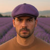 Front-facing portrait of a man wearing a lavender Shetland wool newsboy hat with leather visor in a blooming lavender field in Italy.