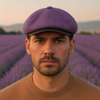Front-facing portrait of a man wearing a lavender Shetland wool newsboy hat with leather visor in a blooming lavender field in Italy.