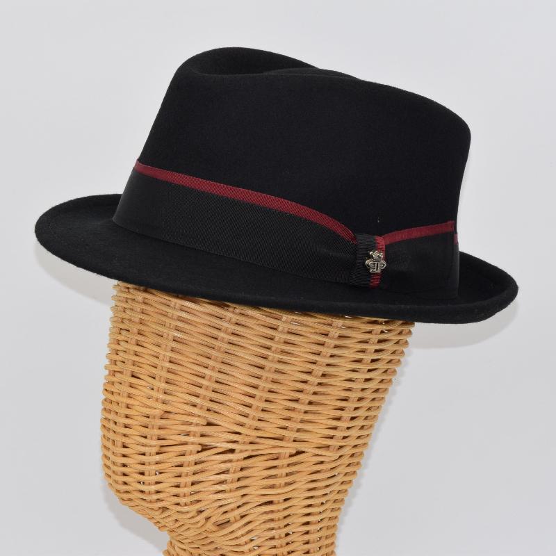 Black alpine hat displayed on a wicker mannequin head, featuring a structured crown, a black ribbon band with a burgundy stripe, and a metallic floral pin detail.