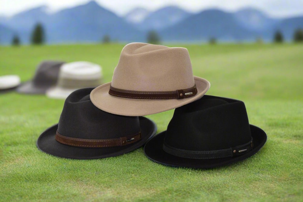 Three alpine hats in beige, brown, and black displayed on a grassy field with a blurred mountain landscape in the background, highlighting their elegant ribbon bands and structured design