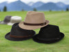 Three alpine hats in beige, brown, and black displayed on a grassy field with a blurred mountain landscape in the background, highlighting their elegant ribbon bands and structured design