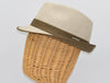 Side view of a beige alpine hat with a structured crown, a brown ribbon band, and a matching brim, displayed on a wicker mannequin head with a metallic logo detail.