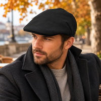 A man wearing a Black Solid Color Coppola Wool Cap , dark charcoal coat, and gray herringbone scarf, photographed outdoors in autumn with blurred trees and warm light.