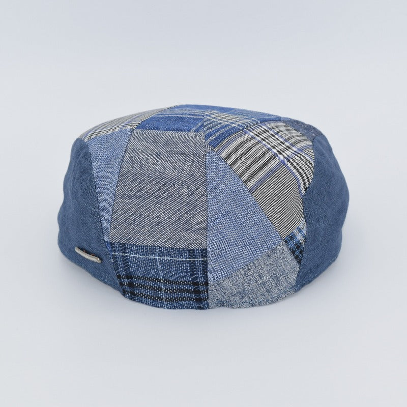 The back view of a blue patchwork driver cap made from an assortment of denim and patterned fabrics, including plaid and herringbone designs. The cap's rounded crown and meticulous stitching create a stylish and contemporary look. A small metallic detail is visible on the side, adding a touch of sophistication.