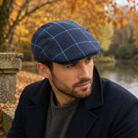 A man wearing a Blue Shetland Wool Fantasy Cap  with a light blue windowpane check pattern, a dark navy coat, and a gray knit sweater, set against an outdoor autumn background with trees and water.
