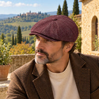 A man wearing a textured Bordo Alba newsboy hat and brown tweed jacket, photographed outdoors against a scenic Tuscan landscape background.