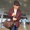 Man in a maroon blazer and flat cap holding a brown leather duffel bag in an airport terminal.