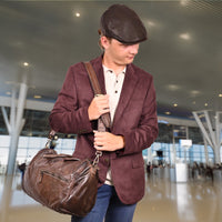 Man in a maroon blazer and flat cap holding a brown leather duffel bag in an airport terminal.