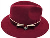 Luxurious burgundy felt fedora with a wide brim, adorned with a velvet band, cream piping, and an elegant bow featuring a decorative metallic emblem, combining classic style with refined detailing.