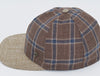 Brown plaid baseball cap with blue and beige checkered patterns, a textured beige brim, and a matching fabric button on top, featuring a small metallic logo detail on the side.