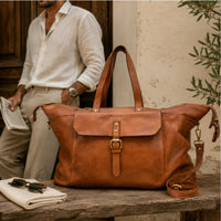 Cognac Santino Leather Travel Bag with a front flap and buckle closure sits on a rustic wooden table. A man in a white shirt and beige trousers stands blurred in the background near a stone wall and greenery.
