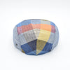 The back view of a multicolored patchwork flat cap showcasing vibrant panels of blue, yellow, orange, and red hues arranged in a geometric design. The herringbone pattern is visible on some panels, adding texture and detail.