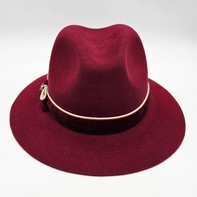 Luxurious burgundy felt fedora with a wide brim, adorned with a velvet band, cream piping, and an elegant bow featuring a decorative metallic emblem, combining classic style with refined detailing.