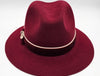 Luxurious burgundy felt fedora with a wide brim, adorned with a velvet band, cream piping, and an elegant bow featuring a decorative metallic emblem, combining classic style with refined detailing.