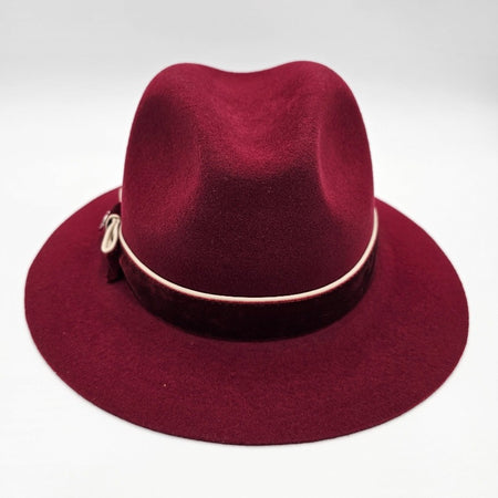 Luxurious burgundy felt fedora with a wide brim, adorned with a velvet band, cream piping, and an elegant bow featuring a decorative metallic emblem, combining classic style with refined detailing.