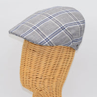 A gray flat cap with a blue and white plaid pattern, displayed on a wicker mannequin head, highlighting its structured side profile and classic style.