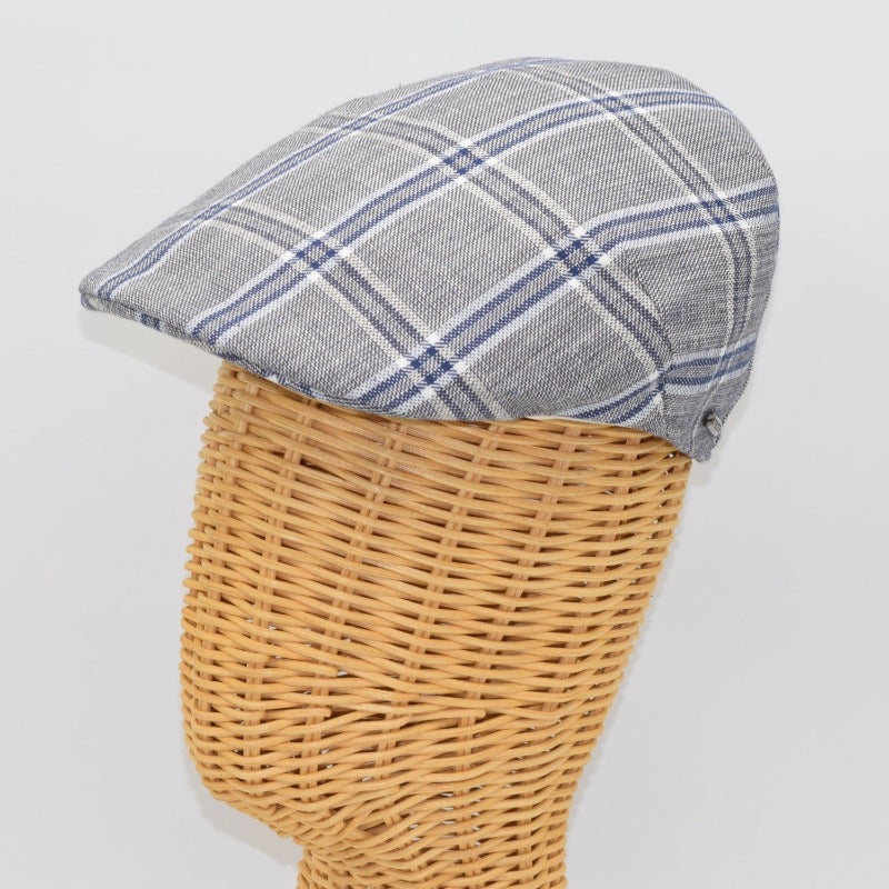 A gray flat cap with a blue and white plaid pattern, displayed on a wicker mannequin head, highlighting its structured side profile and classic style.