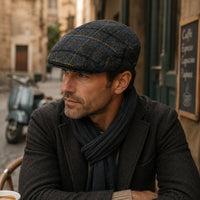 A man wearing a Grey Shetland Wool Cap and navy plaid tweed flat cap, a dark charcoal blazer, and a navy scarf, sitting at an outdoor cafe table with a blurred street and scooter in the background.