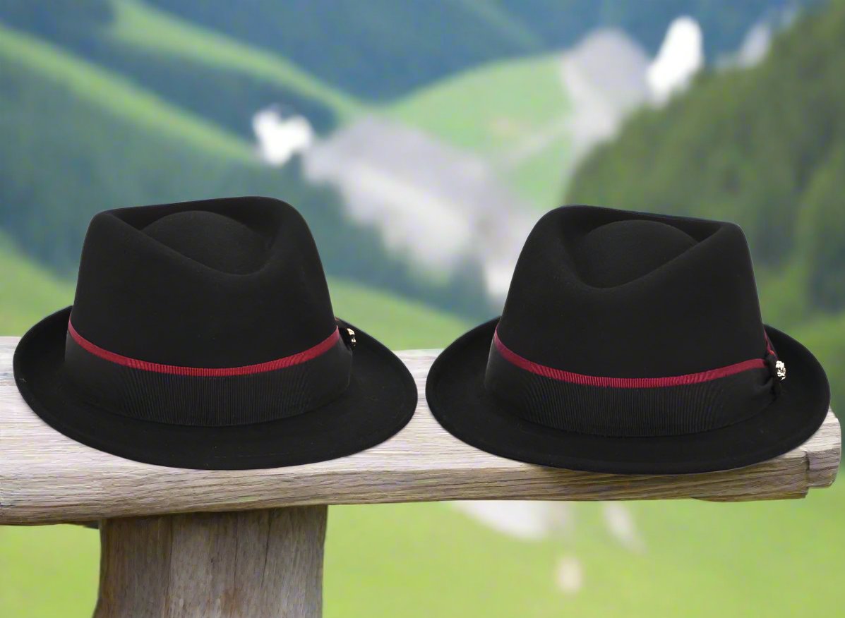 Two black alpine hats with burgundy ribbon bands and metallic pin details displayed on a wooden rail, set against a blurred lush green mountain backdrop.