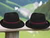 Two black alpine hats with burgundy ribbon bands and metallic pin details displayed on a wooden rail, set against a blurred lush green mountain backdrop.