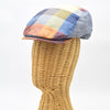 Side view of a vibrant multicolored patchwork flat cap displayed on a wicker mannequin head. The cap features a mix of yellow, blue, orange, and red hues in a checkered pattern, with a slightly curved brim and detailed stitching on the edges.