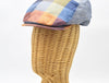 Side view of a vibrant multicolored patchwork flat cap displayed on a wicker mannequin head. The cap features a mix of yellow, blue, orange, and red hues in a checkered pattern, with a slightly curved brim and detailed stitching on the edges.