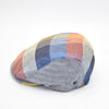 A multicolored patchwork flat cap featuring a vibrant mix of blue, orange, yellow, and red panels with herringbone patterns. The cap has a rounded crown and a small brim, showcasing its artisanal design.