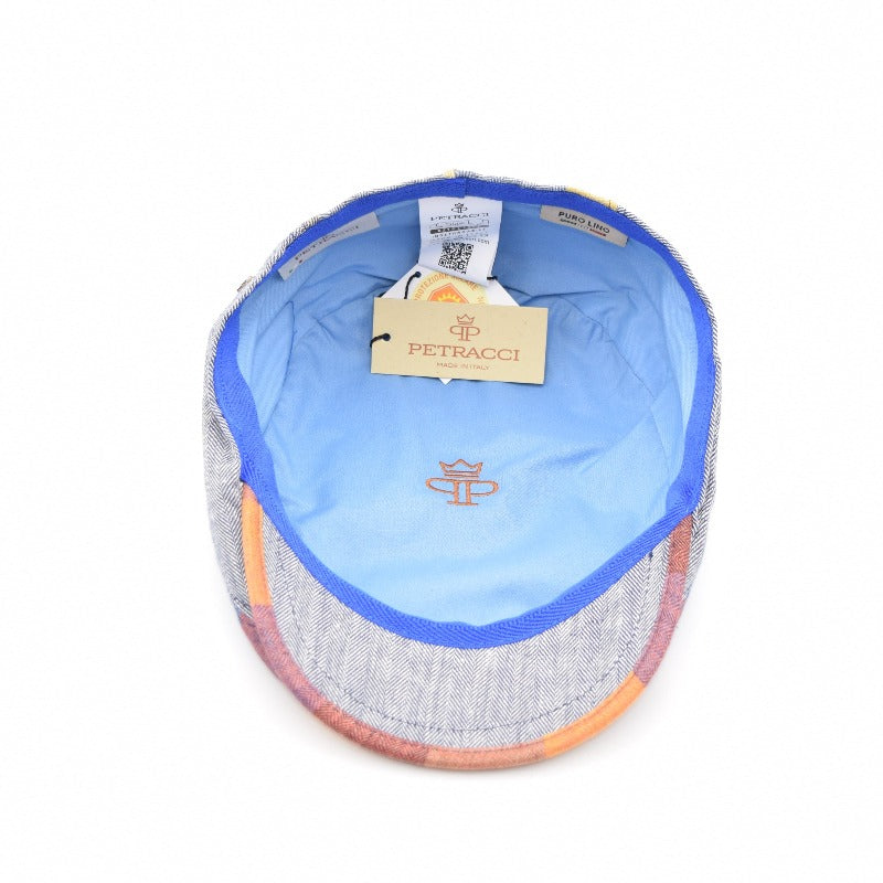  Interior view of a multicolored patchwork flat cap, featuring a light blue lining with an embroidered crown logo in orange. A beige tag with the brand name "Petracci" and "Made in Italy" hangs inside, accompanied by additional labels and a QR code. The cap's edges show hints of its vibrant outer patchwork design.