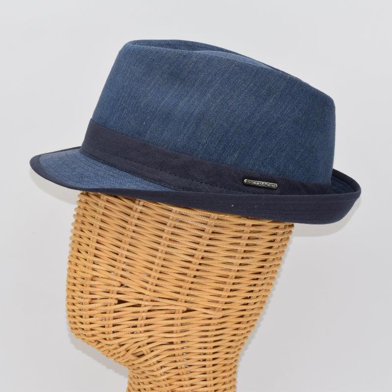 Side view of a blue alpine hat with a structured crown, navy ribbon band, and a small metallic logo detail, displayed on a wicker mannequin head.