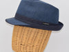 Side view of a blue alpine hat with a structured crown, navy ribbon band, and a small metallic logo detail, displayed on a wicker mannequin head.