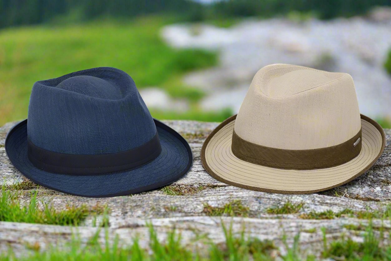 Two alpine hats displayed outdoors on a wooden surface against a blurred grassy and rocky background. One hat is navy with a matching ribbon band, and the other is beige with a brown ribbon band and matching brim.