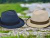 Two alpine hats displayed outdoors on a wooden surface against a blurred grassy and rocky background. One hat is navy with a matching ribbon band, and the other is beige with a brown ribbon band and matching brim.