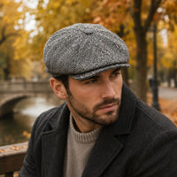 A man wearing a gray and black herringbone patterned Newsboy Fantasy Hat, a dark charcoal coat, and a beige knit sweater, sitting outdoors on a bench with a blurred autumn park background.