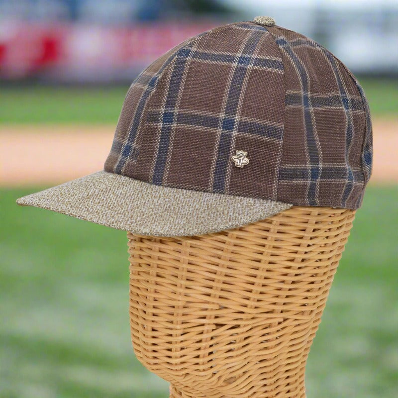 Brown plaid baseball cap with blue and beige checkered patterns, a textured beige brim, and a small metallic logo detail, displayed on a wicker mannequin with a blurred outdoor background.