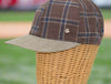 Brown plaid baseball cap with blue and beige checkered patterns, a textured beige brim, and a small metallic logo detail, displayed on a wicker mannequin with a blurred outdoor background.