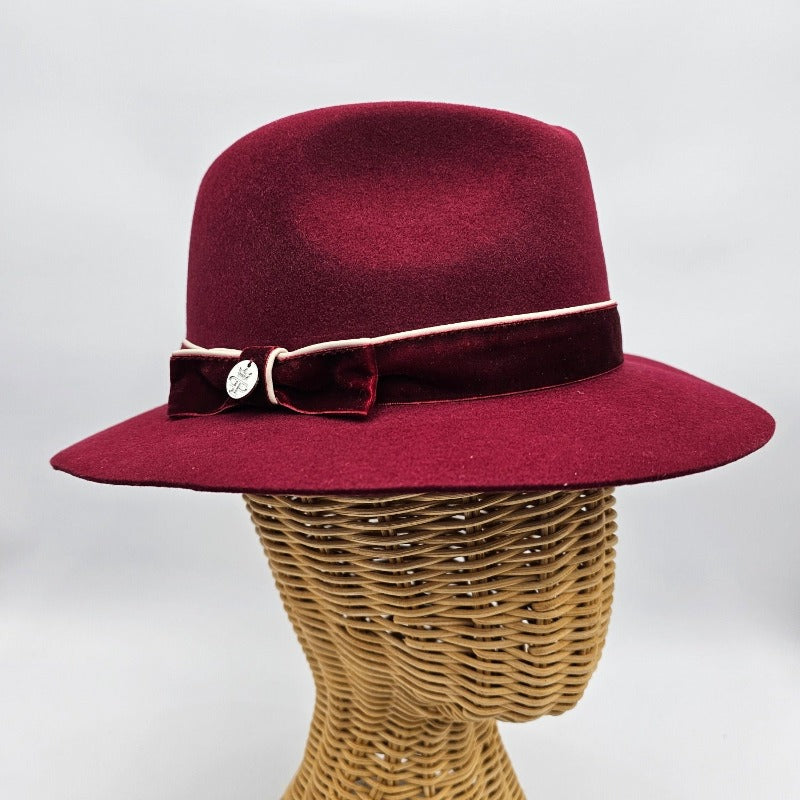 Luxurious burgundy felt fedora with a wide brim, adorned with a velvet band, cream piping, and an elegant bow featuring a decorative metallic emblem, combining classic style with refined detailing.