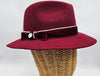 Luxurious burgundy felt fedora with a wide brim, adorned with a velvet band, cream piping, and an elegant bow featuring a decorative metallic emblem, combining classic style with refined detailing.