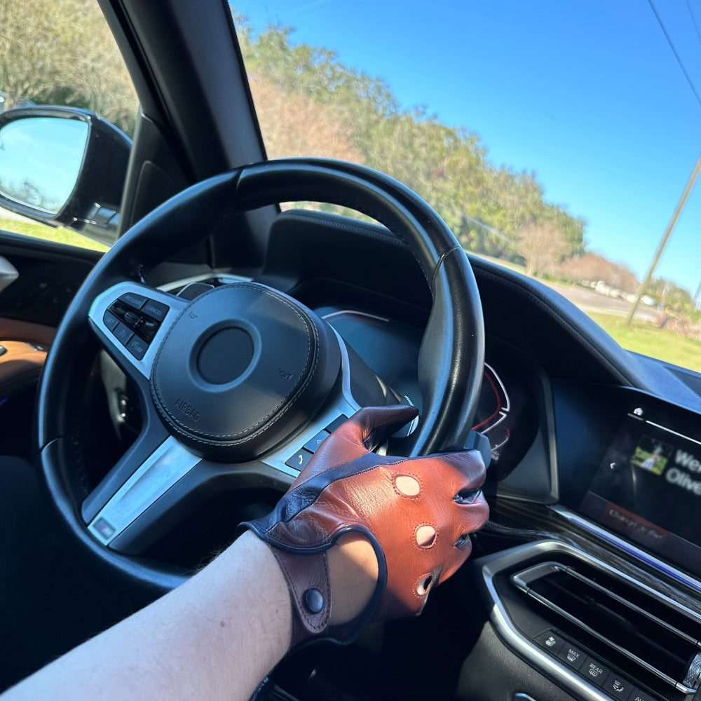 Close-up of a driver wearing two-tone leather driving gloves in brown and deep blue sea, gripping the steering wheel of a car with a modern dashboard, set against a clear blue sky and outdoor scenery.