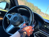 Close-up of a driver wearing two-tone leather driving gloves in brown and deep blue sea, gripping the steering wheel of a car with a modern dashboard, set against a clear blue sky and outdoor scenery.