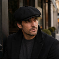 A man wearing a black color Irish Newsboy Shetland Wool Hat and a black coat over a dark gray sweater, photographed outdoors against a softly blurred street background.