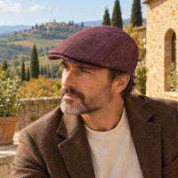 A man wearing a Bordo Alba flat cap and brown blazer, posed outdoors against a scenic Tuscan landscape background.