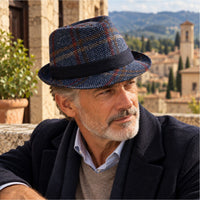 A man wearing a Navy Bruno Wool Trilby Hat with a dark navy band, styled with a dark coat and sweater vest, set against a scenic outdoor background of a stone building and distant hills.