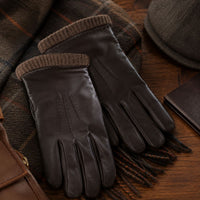 A pair of Brown Men's Lambskin Leather Gloves with ribbed knit cuffs, styled on a brown plaid scarf over a wooden surface with a hat and leather accessories nearby.