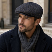A man wearing a Multicolor Perugia Newsboy Hat , a dark navy wool coat, and a gray knit scarf, photographed in an outdoor urban setting.