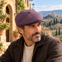 A man wearing a Lavender Norcia Newsboy hat and a brown textured jacket, set against a scenic background of a stone building and a Tuscan hillside village.