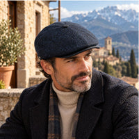 A man wearing a dark navy Norcia Newsboy Hat and a dark herringbone jacket over a beige turtleneck and plaid scarf, set against a scenic background of a stone building and snow-capped mountains.
