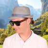 Man wearing a grey alpine hat and sunglasses with a mountainous landscape in the background