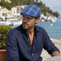 A man wearing a blue portofino flat cap and a navy blue button-down shirt, seated outdoors with a marina and coastal town in the background.